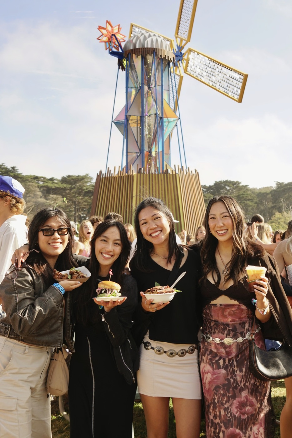four friends holding festival food in front of a glittering windmill sculpture